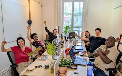 People at a long shared desk with laptops and plants raise their arms and cheer toward the camera.