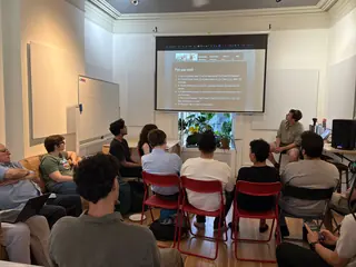 A full room seen from behind: people on red chairs and a sofa face a projected slide.