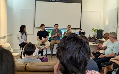 Four panelists on stools under a projection screen face an audience seated on a sofa and chairs.