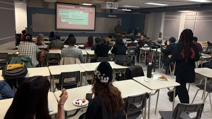 A lecture room seen from the back: people seated at tables face a projected slide and the speaker beside it.